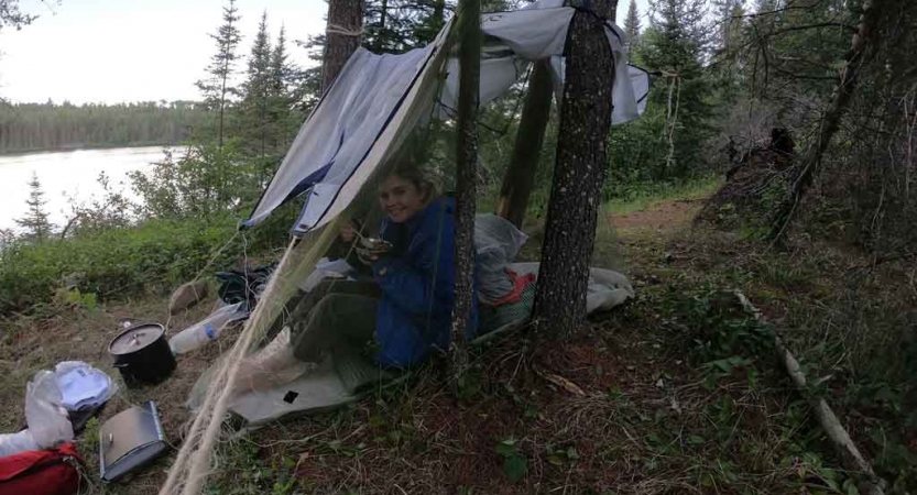 a student smiles from a tarp shelter in a wooded area beside a body of water 